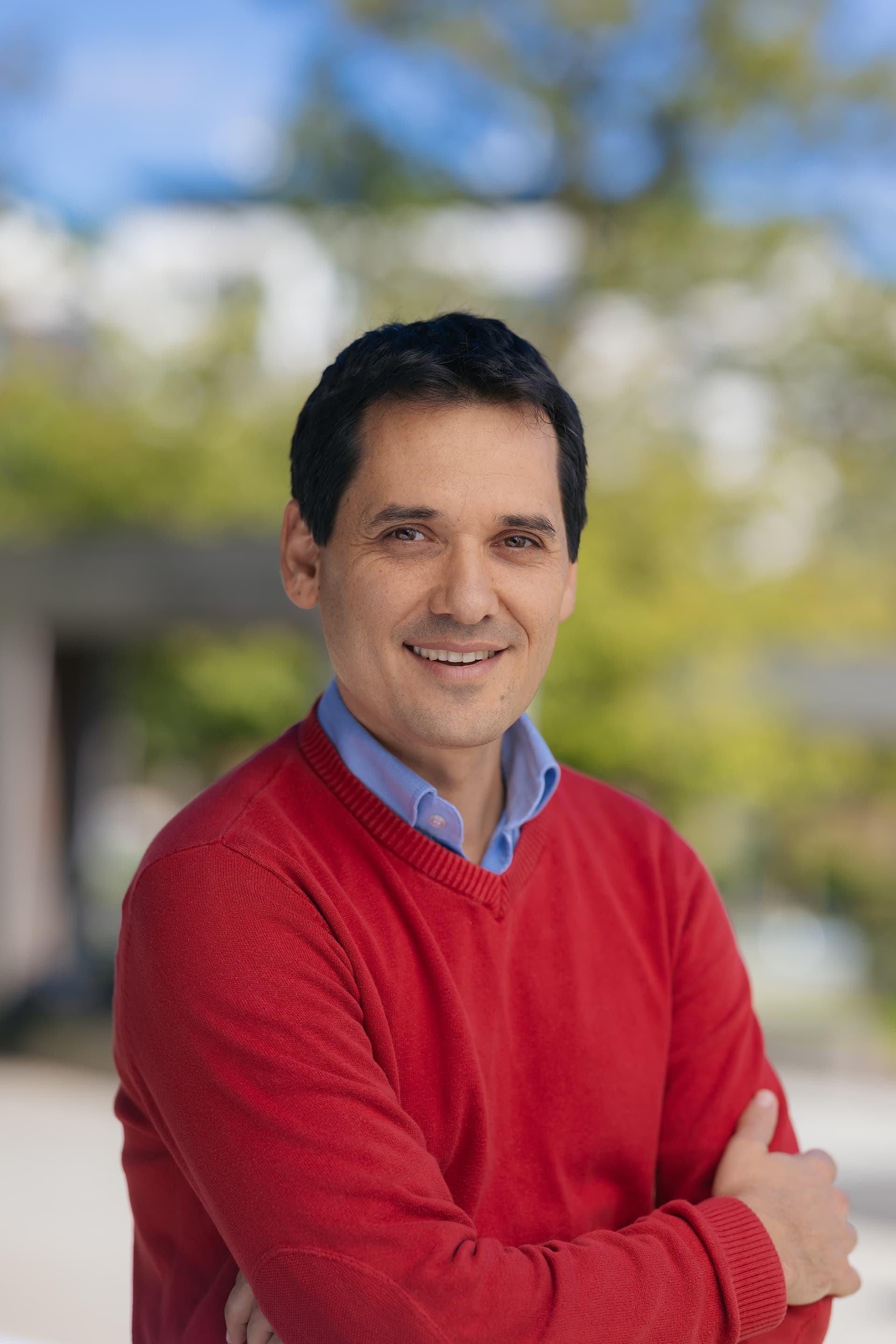 Smiling man wearing a red sweater and blue shirt standing outdoors with a blurred green background.